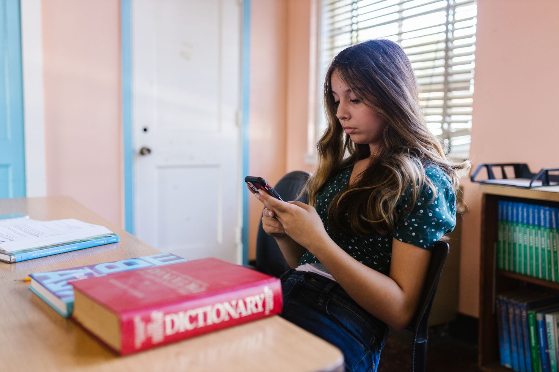 Estudante lendo no celular com o dicionário fechado sobre a mesa